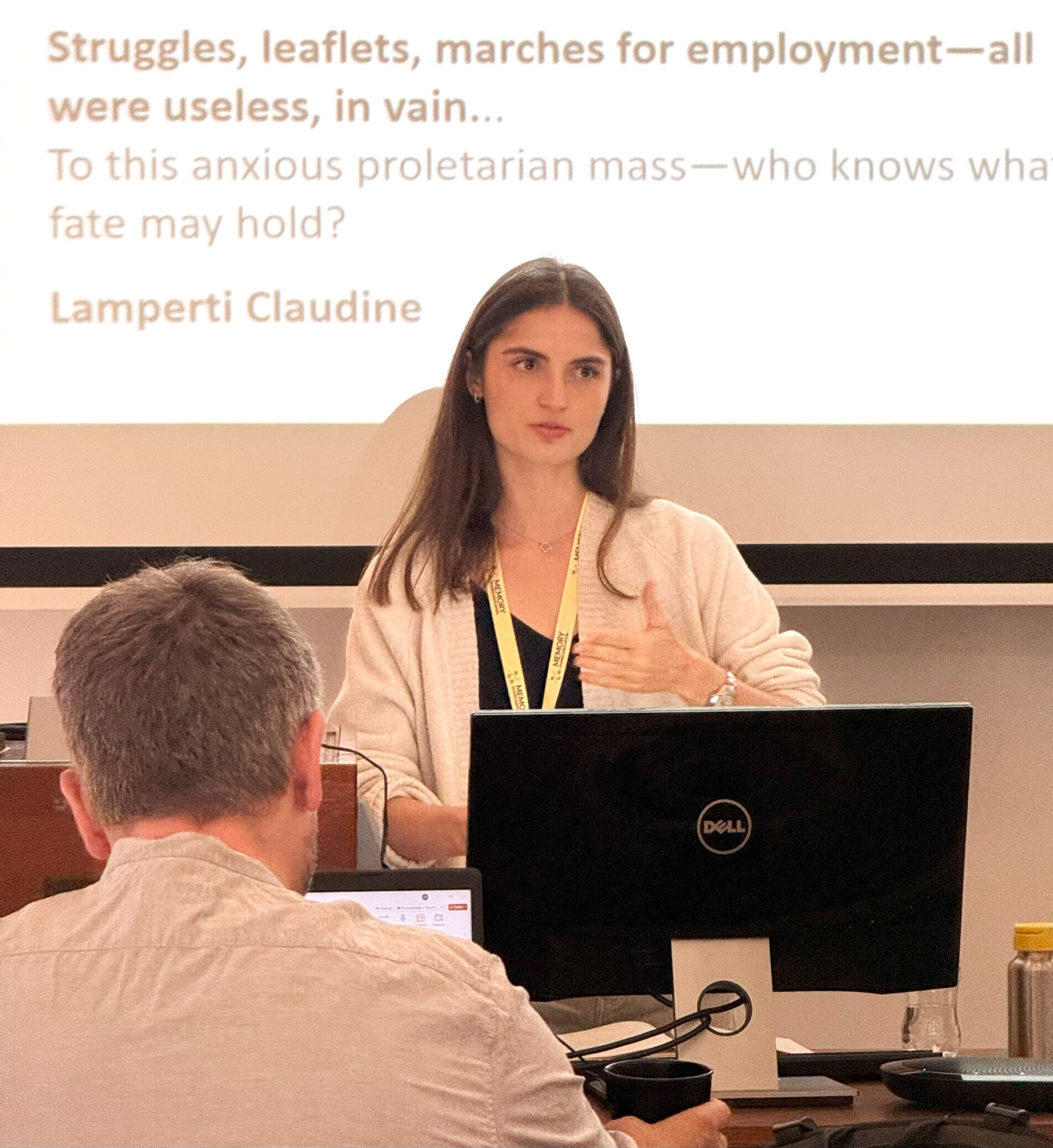 cholar Zoe stood in front of a laptop and presenting, with presentation slides showing text behind her.